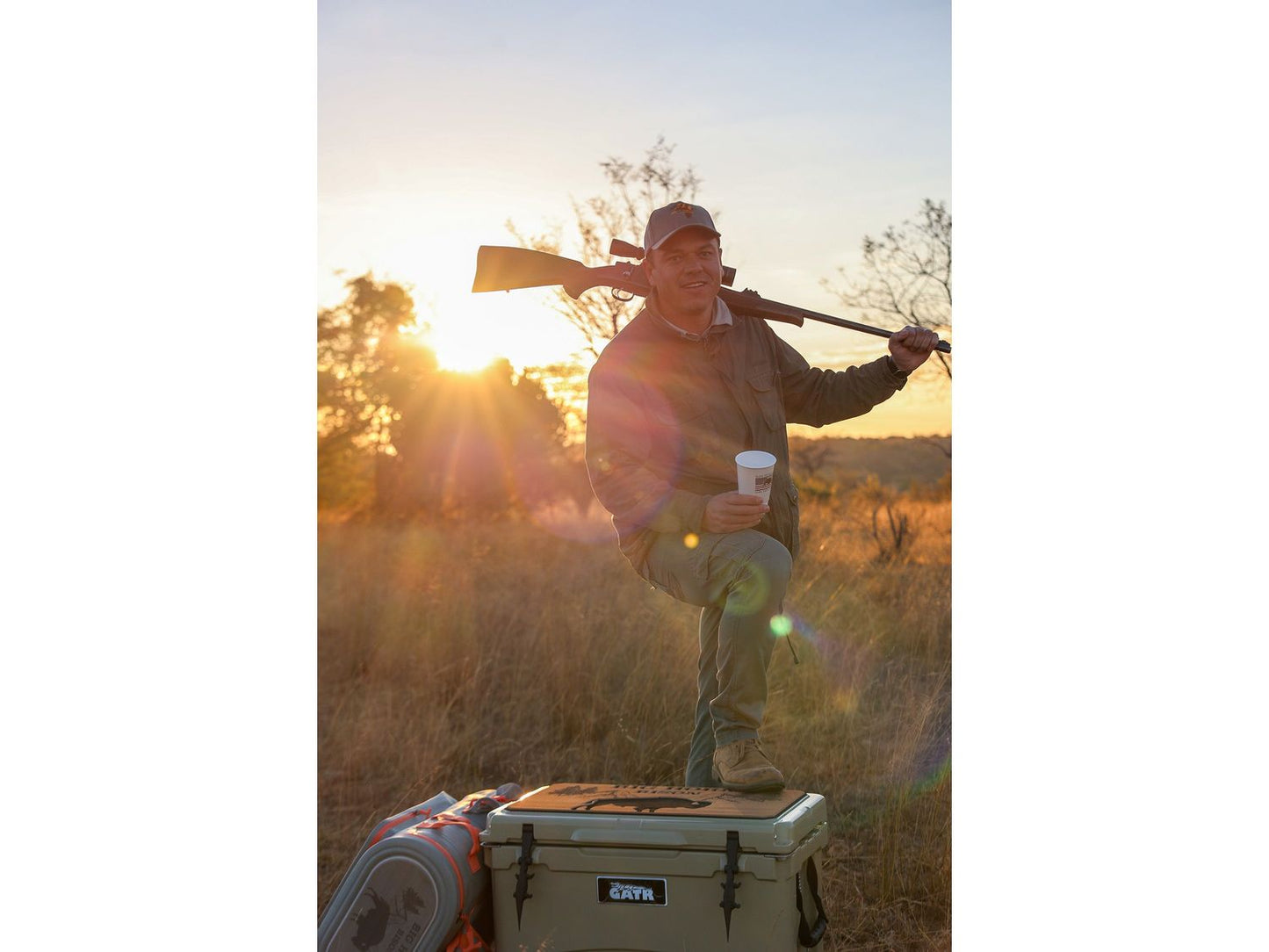 Man in a field with a rifle over his shoulder, holding a coffee cup, standing on a cooler.